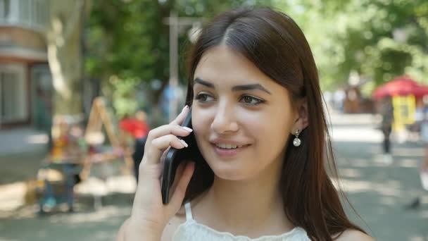 Young woman is chatting on her phone smiling in a street in summer in slo-mo                              Portrait of a cheery young woman with sparkling eyes and long loose hair talking on her smartphone in a green street in summer in slow motion - Séquence, vidéo