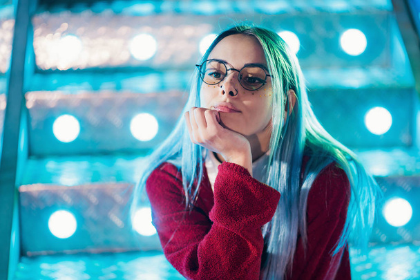 Hipster girl with blue dyed hair and sequins as freckles. Woman in red clothing and nose piercing, transparent glasses, ears tunnels, unusual hairstyle in amusement park - Foto, Bild
