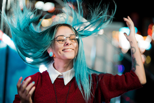 Hipster girl waving head in different directions, playing with dyed blue hair. Woman with nose piercing, transparent glasses, ears tunnels, unusual hairstyle in amusement park - Valokuva, kuva