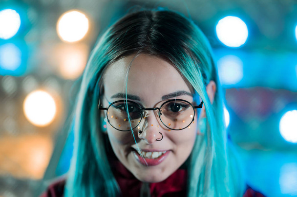 Hipster girl with blue dyed hair and golden sequins as freckles. Woman with nose piercing, transparent glasses, ears tunnels, unusual hairstyle stands in amusement night park. - Photo, Image