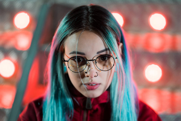 Hipster girl with blue dyed hair and golden sequins as freckles. Woman with nose piercing, transparent glasses, ears tunnels, unusual hairstyle stands in amusement night park. - Photo, image