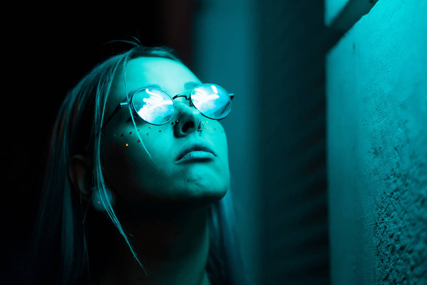 Millennial enigmatic pretty girl with unusual dyed hairstyle near glowing neon wall at night. Blue hair, golden sequins as freckles,nose piercing. Mysterious hipster teenager in glasses - Fotó, kép