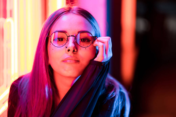 Millennial enigmatic pretty girl with unusual dyed hairstyle near glowing neon wall at night. Blue hair, golden sequins as freckles,nose piercing. Mysterious hipster teenager in glasses - Photo, image