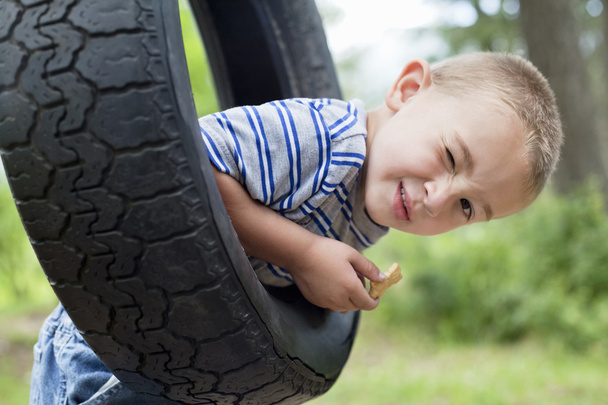Portrait of a young boy winking while swinging on tire - 写真・画像