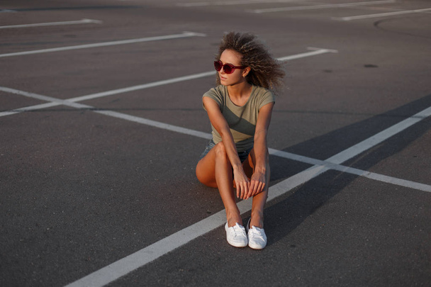 stylische junge Hipster-Model Frau mit lockiger Frisur in Sonnenschutzbrille in Jeanshosen mit stylischem T-Shirt und weißen Turnschuhen auf dem Asphalt auf der Straße sitzend - Foto, Bild