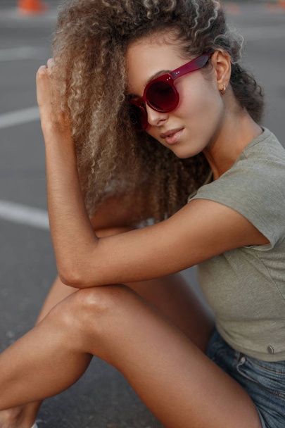 Hipster donna alla moda con i capelli ricci con eleganti occhiali da sole rosa in abiti di moda seduti sulla strada
 - Foto, immagini