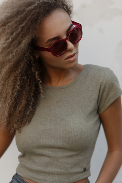 Portrait of a beautiful fashionable young curly woman with pink sunglasses in a stylish T-shirt on the street - Fotó, kép