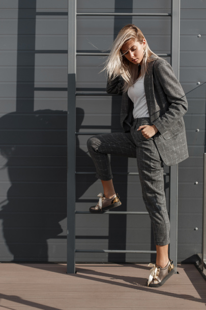 Jovem mulher modelo bonita em terno de moda vintage posando perto da parede com escada no dia ensolarado
 - Foto, Imagem