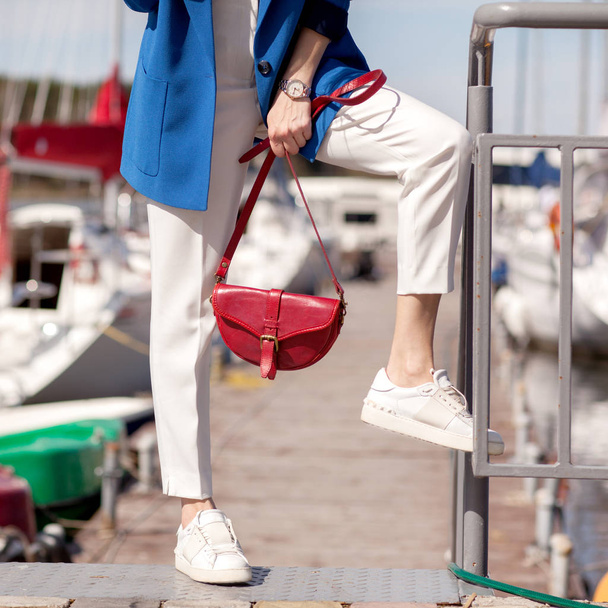 Straat, heldere stijl. Jonge vrouw in een blauwe jas, witte sneakers, witte broek, een rode handtas, Sunglassesand en een rode case voor zonnebril. Details. Sguare afbeelding foto - Foto, afbeelding
