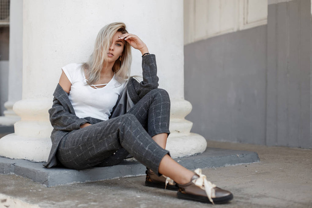 Fashionable beautiful stylish woman in a gray coat and stylish pants with shoes sitting near a white column on the street - Photo, Image