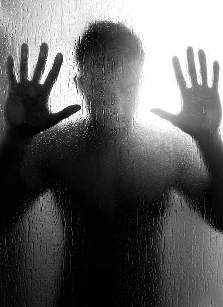 homme nu dans la douche tenant la main contre la paroi de verre, un signe de dépression, de solitude et de tristesse. Sa main repose sur une cloison de verre avec une goutte d'eau
. - Photo, image