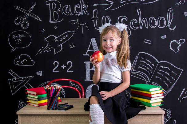 Cheerful schoolgirl sitting on the desk with books, school supplies, holding a red apple in the right hand - Fotó, kép