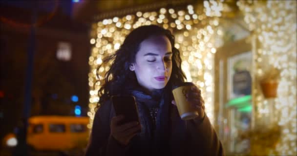 Attractive woman using mobile phone while walking through the streets on the background of festive lights of night city. - Felvétel, videó