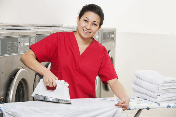 Portrait of a happy woman employee in red uniform ironing clothes in Laundromat - Photo, image
