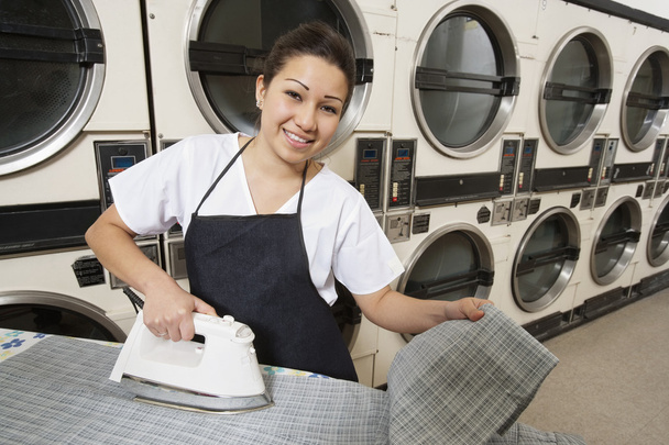 Portrait of a happy woman wearing apron ironing in front of washing machines - Photo, image