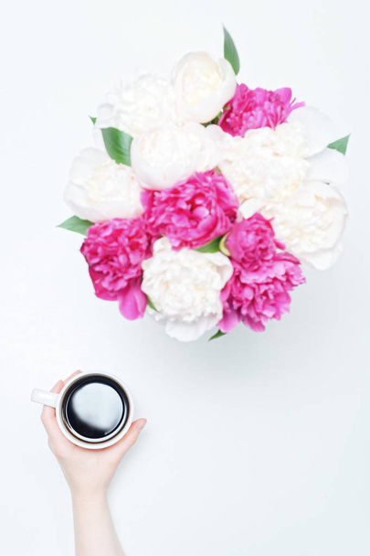 Working place with hand holding cup of coffee and white and pink peony flowers on white table background. Flat lay, top view - Фото, зображення
