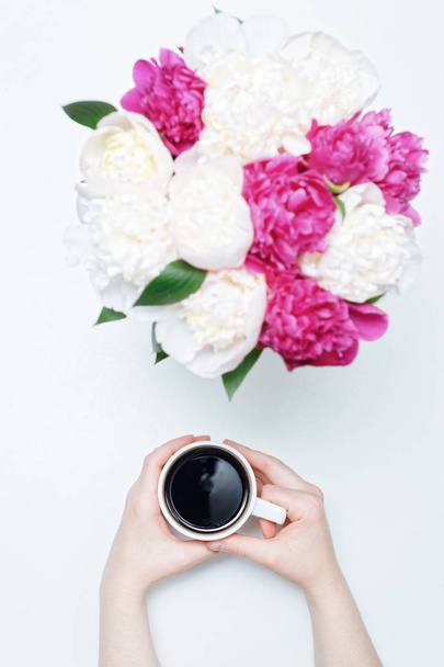 Werkplaats met hand holding kopje koffie en kleurrijke witte en roze pioenroos bloemen op witte tafel achtergrond. Vlakke lay, bovenaanzicht - Foto, afbeelding