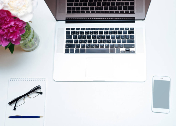Arbeitsplatz mit Laptop, Brille, Notizbuch, Stift und rosa und weißen Pfingstrosenblüten auf dem weißen Tischhintergrund. flache Lage und Draufsicht - Foto, Bild