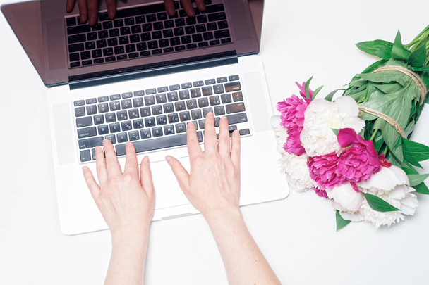 Arbeitsplatz mit Laptop, Frauenhänden, Notizbuch und rosafarbenen und weißen Pfingstrosenblüten auf dem weißen Tischhintergrund. flache Lage, Draufsicht - Foto, Bild