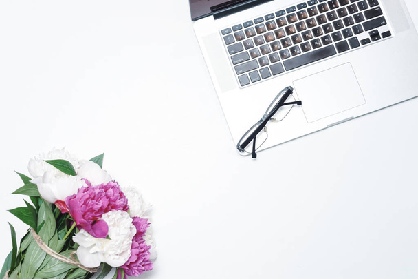 Lugar de trabajo con portátil, gafas y con flores de peonía rosa y blanca sobre el fondo de la mesa blanca. Piso tendido, vista superior
 - Foto, imagen