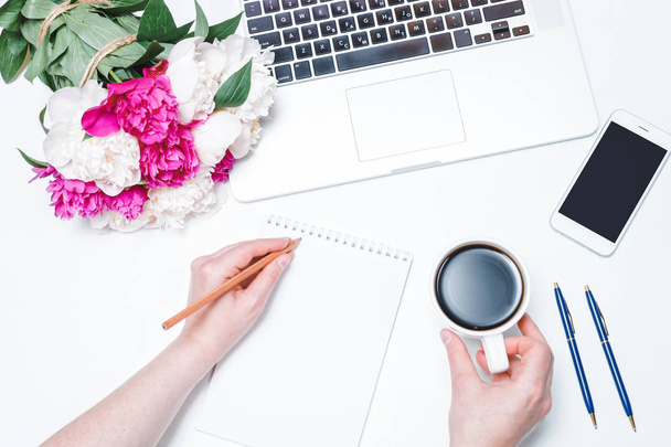 Workspace with mobile phone, laptop, cup of coffee and female hands, notebook, pen and pink and white peony flowers on the white background. Flat lay and top view. Freelancer working place - Valokuva, kuva