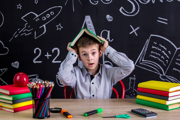 Scared and puzzled schoolboy sitting at the desk holding the book above the head, surrounded with school supplies. Chalkboard as a background - Photo, Image