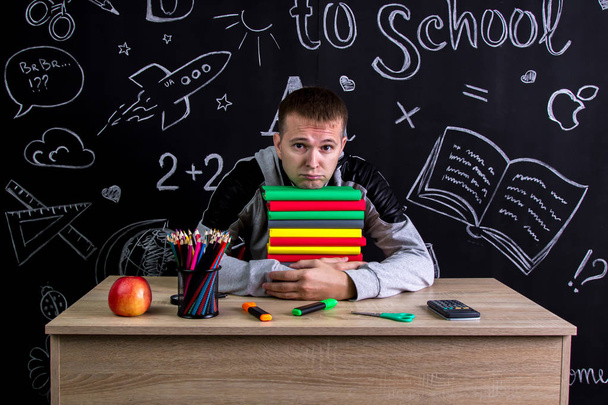 Unhappy youngster sitting at the desk with a pile of books under the chin, surrounded with school supplies. Chalkboard as a background - Photo, Image