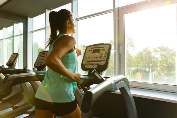 Side view of Young concentrated sports woman running on treadmill near the window in gym - Photo, Image