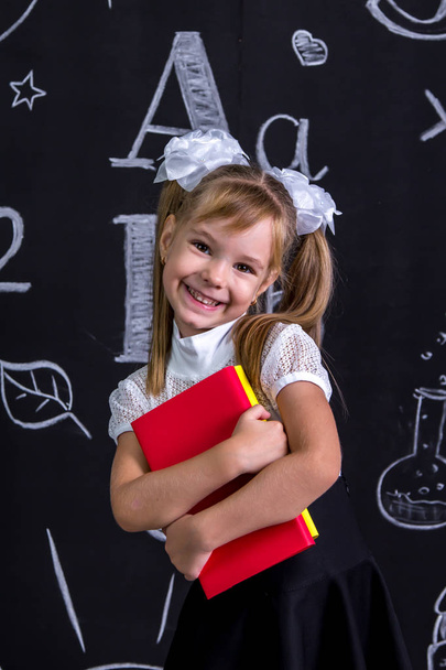 Lächelndes, glückliches Schulmädchen, das kokett vor der Tafel steht, als Hintergrund ein paar Bücher, die sie umarmen. Porträtbild - Foto, Bild