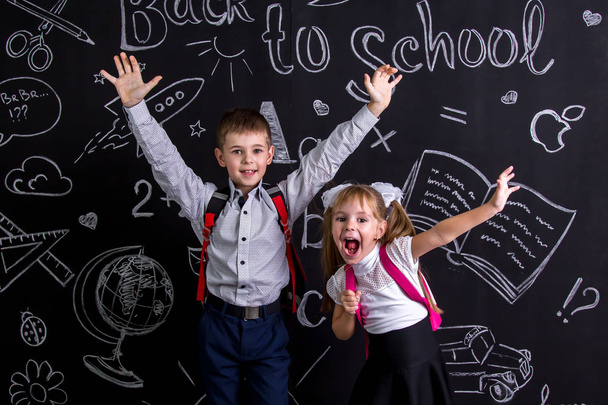 Podekscytowany i wesoły schoolkids stojąc przed chalkboard jako tło z plecakiem na plecach Wyświetlono szczęśliwy emosions za pomocą podniesione ręce - Zdjęcie, obraz