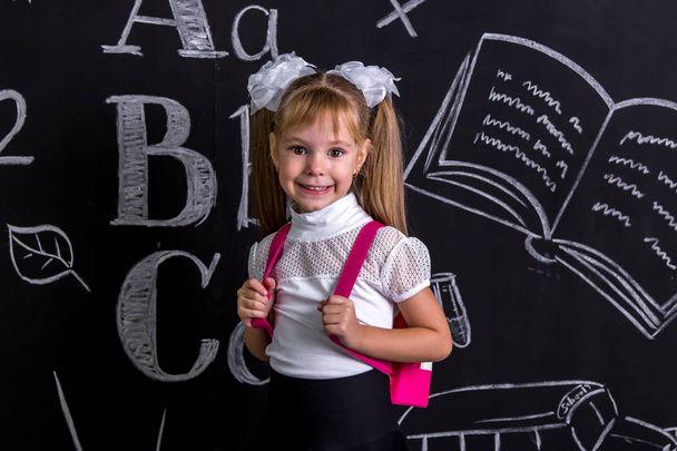 Lächelndes Schulmädchen, das mit einem Rucksack auf dem Rücken vor der Tafel steht. vergrößerte Ansicht - Foto, Bild