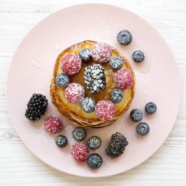 Pancakes with berries, honey and sugar on a pink plate on a white wooden table, overhead view. Flat lay, top view, from above. Close-up. - Photo, Image