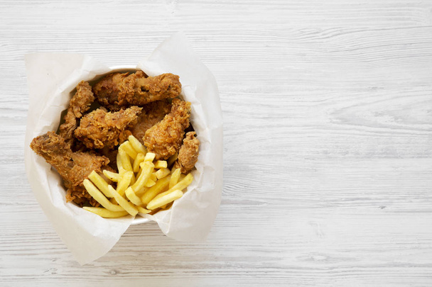 Tasty fast food: fried chicken drumsticks, spicy wings, French fries and tender strips in paper box over white wooden background, view from above. Flat lay, top view, from above. Space for text. - Foto, Imagen