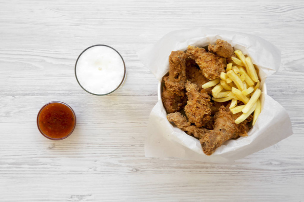 Fried chicken drumsticks, spicy wings, French fries and tender strips in paper box, sauce and cold beer over white wooden background, overhead view. Flat lay, top view, from above.  - Photo, image