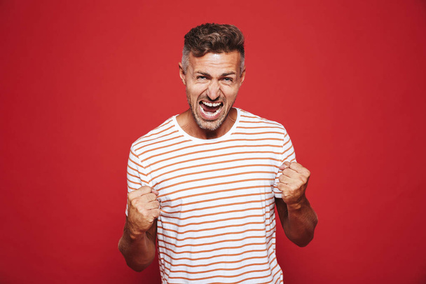 Emotional furious man in striped t-shirt screaming and showing fists isolated over red background - Foto, immagini