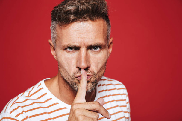 Serious man in striped t-shirt holding index finger on lips with strict look isolated over red background - Zdjęcie, obraz