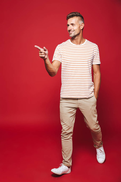 Full length photo of young man in striped t-shirt gesturing index fingers aside at copyspace isolated over red background - Foto, Bild