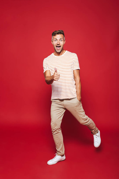 Full length photo of attractive man in striped t-shirt smiling and showing thumb up isolated over red background - Fotografie, Obrázek