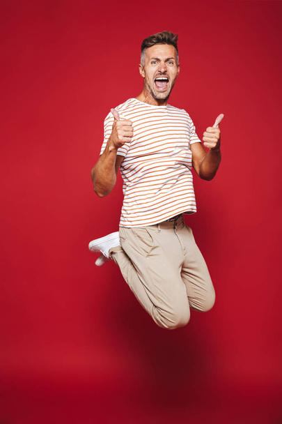 Full length photo of happy man in striped t-shirt jumping and showing thumbs up isolated over red background - Фото, зображення