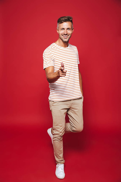 Full length photo of positive man in striped t-shirt smiling and showing thumb up isolated over red background - Photo, Image