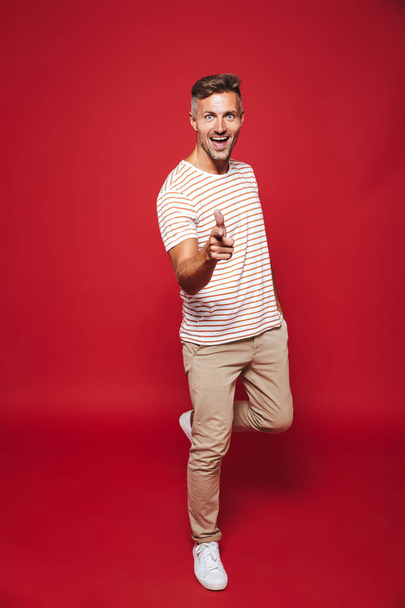 Full length photo of unshaved man in striped t-shirt smiling and showing thumb up isolated over red background - Фото, изображение