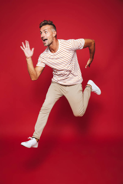 Full length photo of caucasian man in striped t-shirt running and jumping isolated over red background - Zdjęcie, obraz