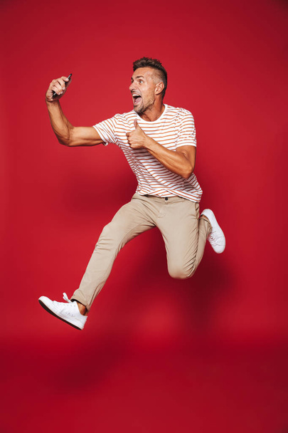 Full length photo of caucasian man in striped t-shirt jumping and taking selfie on smartphone isolated over red background - Foto, Bild