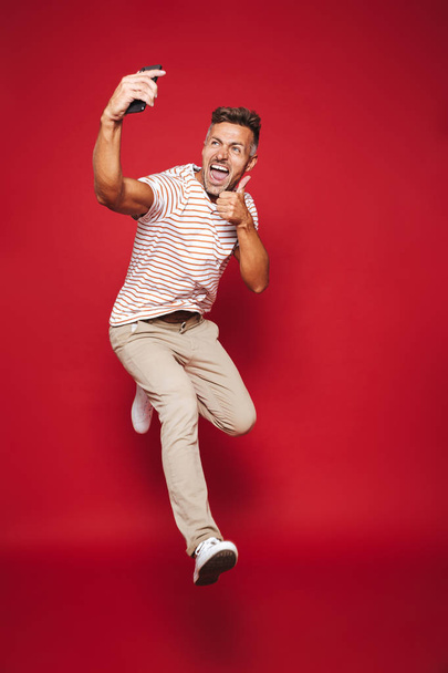 Full length photo of caucasian man in striped t-shirt jumping and taking selfie on mobile phone isolated over red background - Foto, Bild