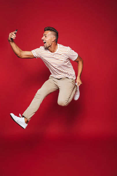 Full length photo of optimistic man in striped t-shirt jumping and taking selfie on smartphone isolated over red background - Foto, imagen