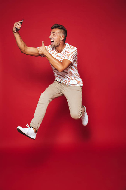 Full length photo of handsome man in striped t-shirt jumping and taking selfie on smartphone isolated over red background - Photo, image