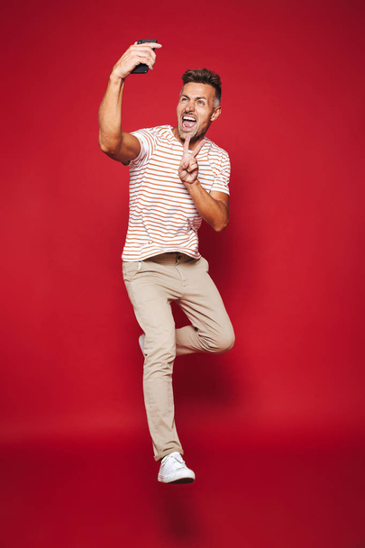 Full length photo of brunette man in striped t-shirt jumping and taking selfie on smartphone isolated over red background - Fotó, kép