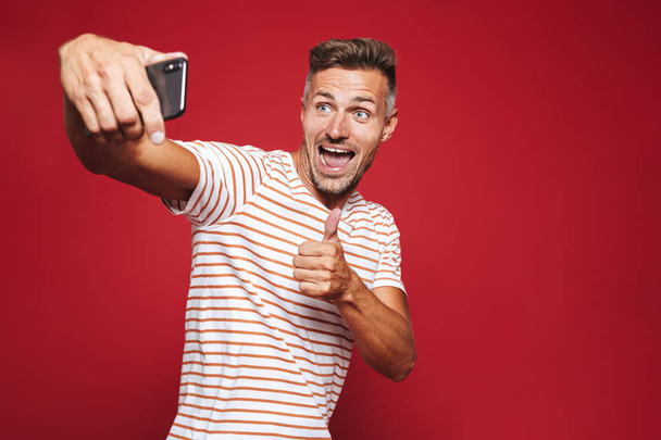 Portrait of a cheerful man standing over red background, taking a selfie, giving thumbs up - Foto, Imagen