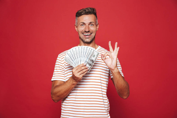 Image of rich guy in striped t-shirt smiling and holding fan of money banknotes isolated over red background - Φωτογραφία, εικόνα