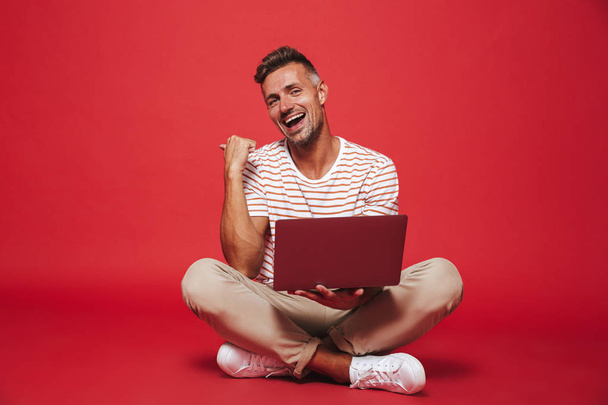 Image of handsome man 30s in striped t-shirt smiling and using laptop while sitting on floor with legs crossed isolated over red background - Photo, Image